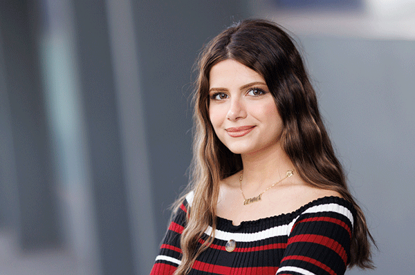 Gemma from Syria looks at the camera. She's wearing a red, black and white dress and her brown hair hangs loose over her shoulders. 