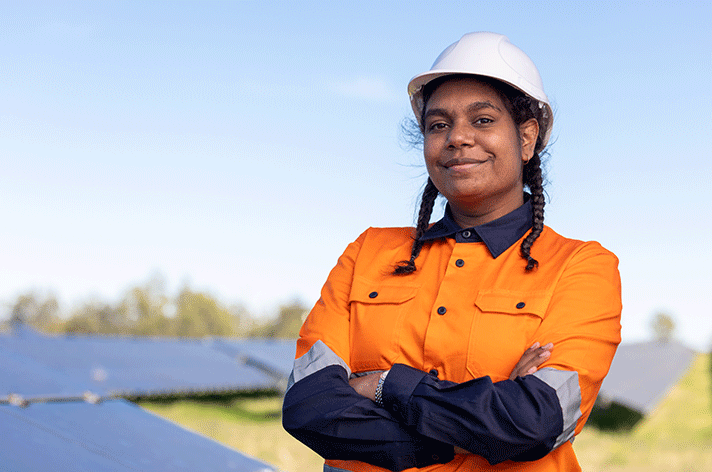 Hero image of the MAX RAP 2022. A young Indigenous woman is in high vis workwear including a safety helmet and stands upon a roof overlooking other roofs with solar panels.