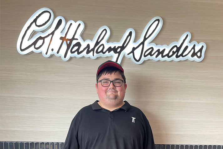 Smiling Shaun stands in front of a KFC sign, "Col. Harland Sanders" in cursive script