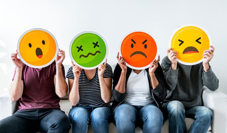 people sat ona  bench holding up cardboard emojis showing different emotions like sad, angry, shocked