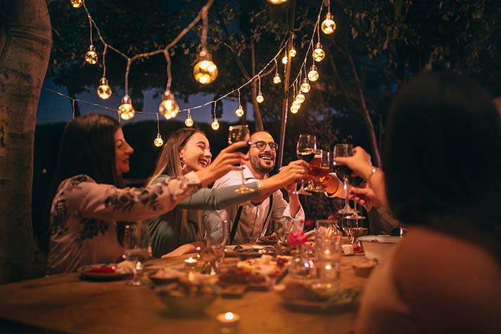 A group of adults "cheers" with alcoholic drinks at a dinner table at night set under festoon lights. 